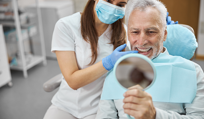 Older man sitting in a dental chair looking in a handheld mirror at his new dental implants from Frank Dunlap, DDS in Houston, TX